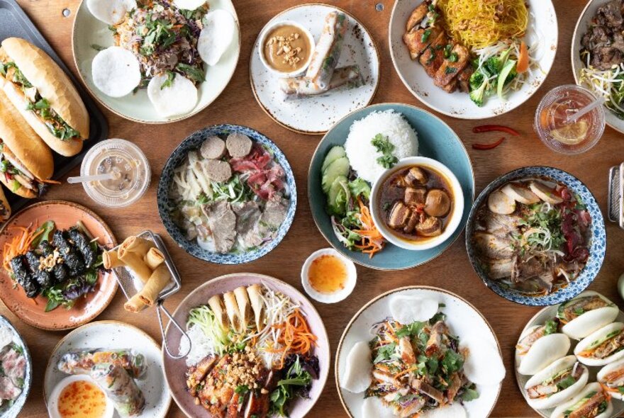 Overhead view of many Vietnamese dishes of food on a wooden table, including sauces and condiments, banh mi and rice.