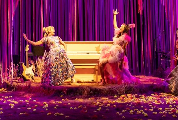 Two actors performing on a stage with purple curtains and a piano.