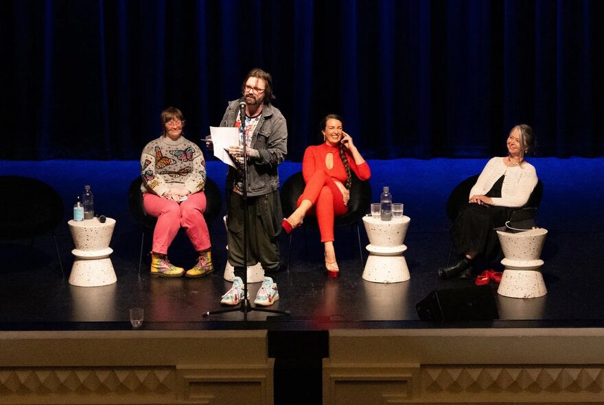 A panel of people on stage, three seated and one standing up and talking into a microphone.