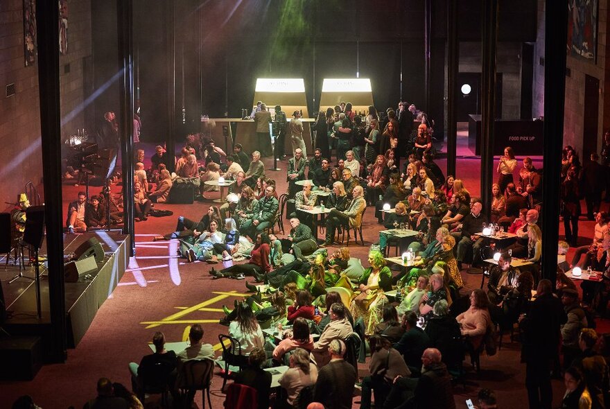 The Great Hall at NGV venue with a band performing on the stage in front of large audience seated at small tables and chairs, some standing near the bar towards the rear of the space.