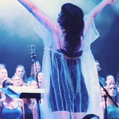 A back-view shot of a female choir conductor leading a large choir performance on stage.