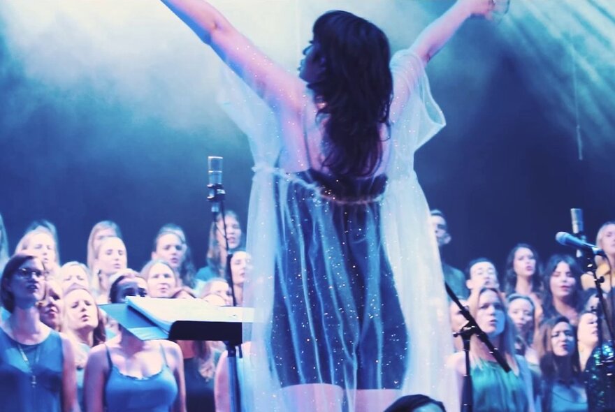 A back-view shot of a female choir conductor leading a large choir performance on stage.
