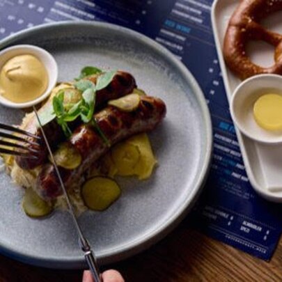 A dish of German sausages, mash, sauerkraut and mustard, next to a pretzel with butter.