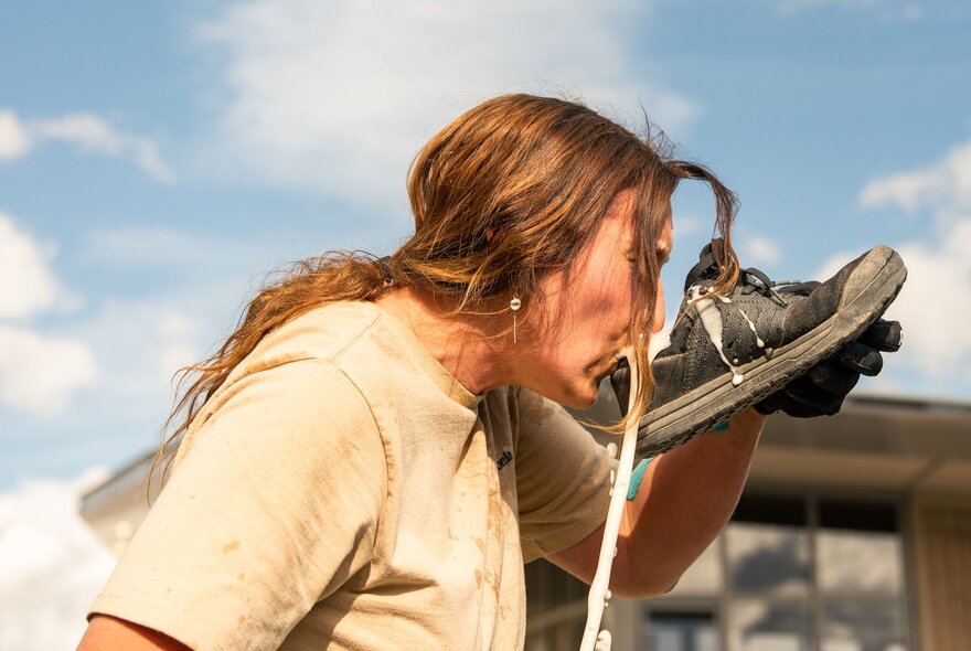 Profile of a person with long hair wearing a t-shirt, holding a well-worn running shoe or sneaker up to their lips, as if drinking liquid from it.