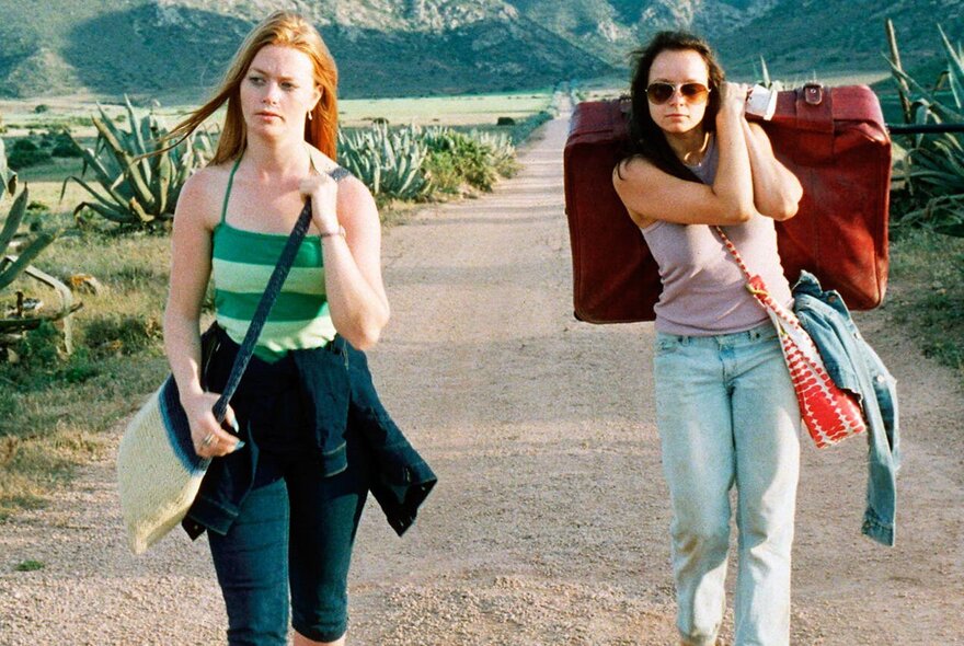 A still image from a film showing two woman walking along a road in a remote location, one carrying a large suitcase over her shoulder. 