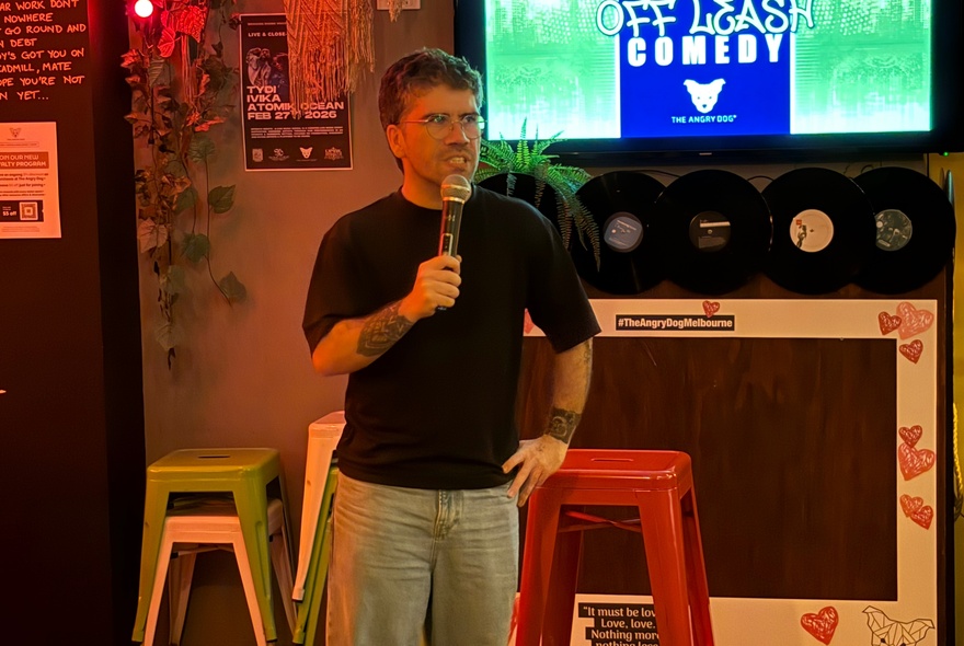 A man in a black t-shirt standing with a microphone in front of a presentation screen and two stools, inside a bar setting. 