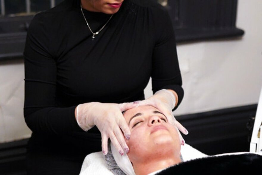 A woman receiving a facial treatment from a facialist wearing black, with her gloved hands massaging the woman's forehead.