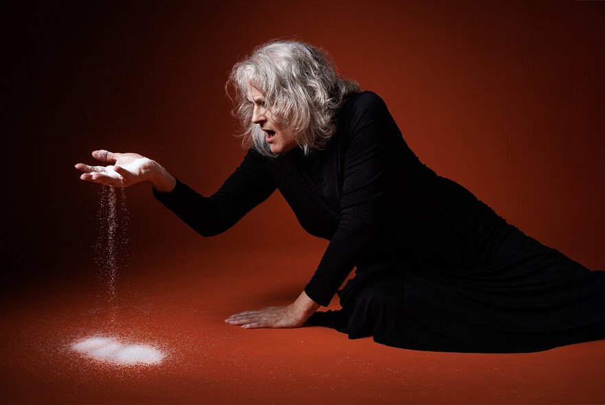 Actor with grey hair reclining in a red space, pouring white sand through her fingers and grimacing.