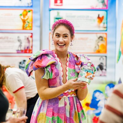 A woman at a food show dressed in a colourful dress and presenting a product to onlookers. 