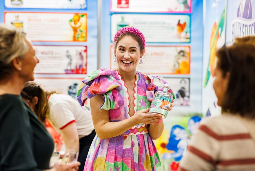 A woman at a food show dressed in a colourful dress and presenting a product to onlookers. 