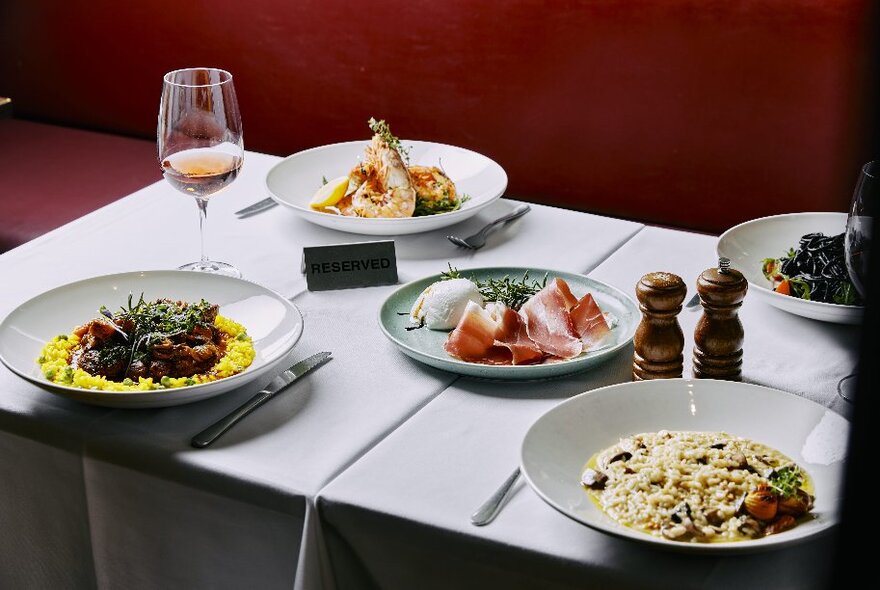 A table set with white linen in a restaurant, set with five dishes, and glasses of wine.