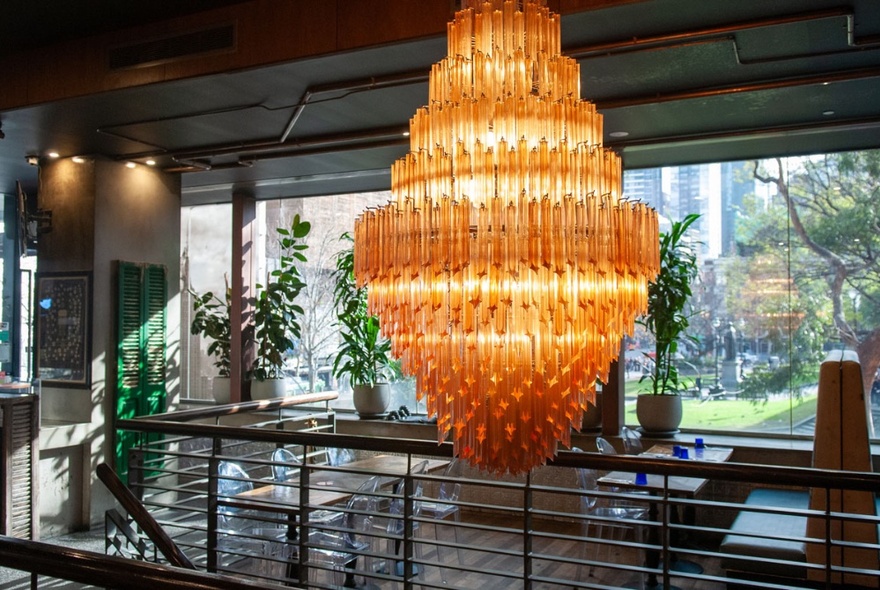 A large orange chandelier hanging above the staircase inside the venue Father's Office, with the grassy lawns in front of the State Library visible through the balcony windows.