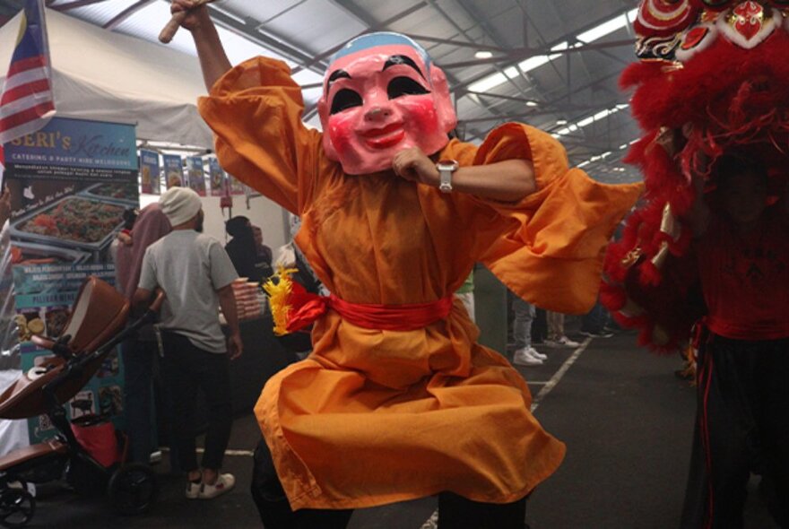 Malaysian costume character wearing orange robes and a pink mask, dancing in an undercover space.