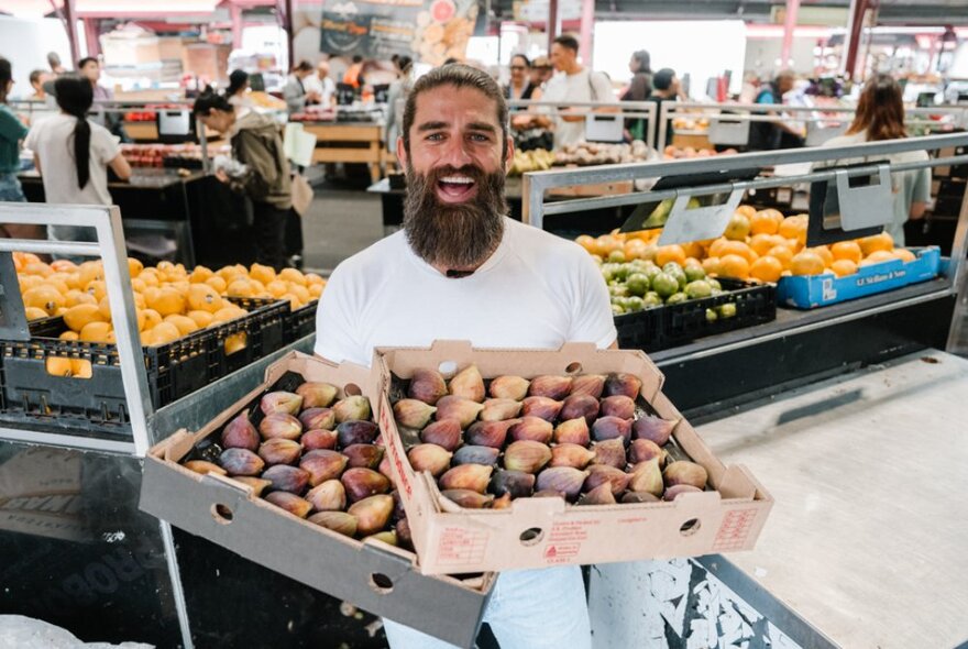 Bearded man holding two trays of ripe figs in a fruit and veg market.