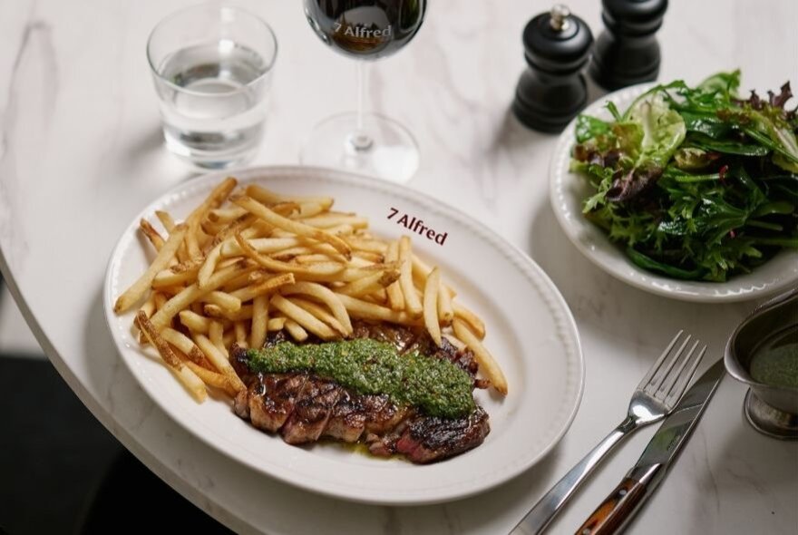 Steak frites with a chimichuri sauce on a white plate, accompanied by a green salad in a separate dish.