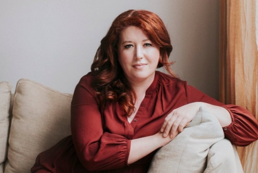Author Jane Harper with her auburn hair arranged over one shoulder, leaning on the arm of a neutral coloured sofa. 