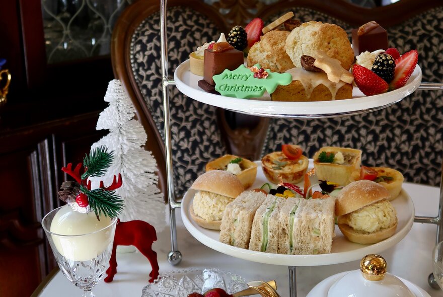 Tiered afternoon tea stand with sandwiches, scones and cakaes, on a table decorated with Christmas deer and tree.