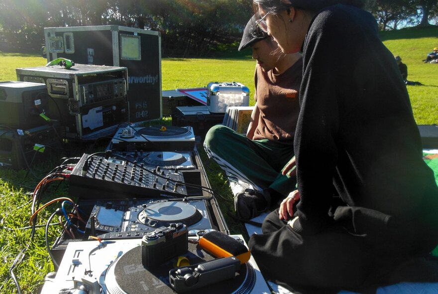 Two people sitting on grass in a large open park, musical equipment, a turntable and consoles in front of them.