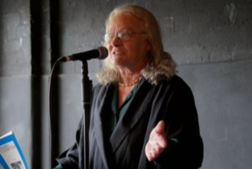 A woman with long, light-colored hair and glasses, wearing a dark jacket speaks into a microphone on a stand.