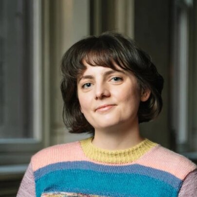 Portrait of a subtly smiling young woman with short brown hair, wearing a pink and blue striped knitted jumper.