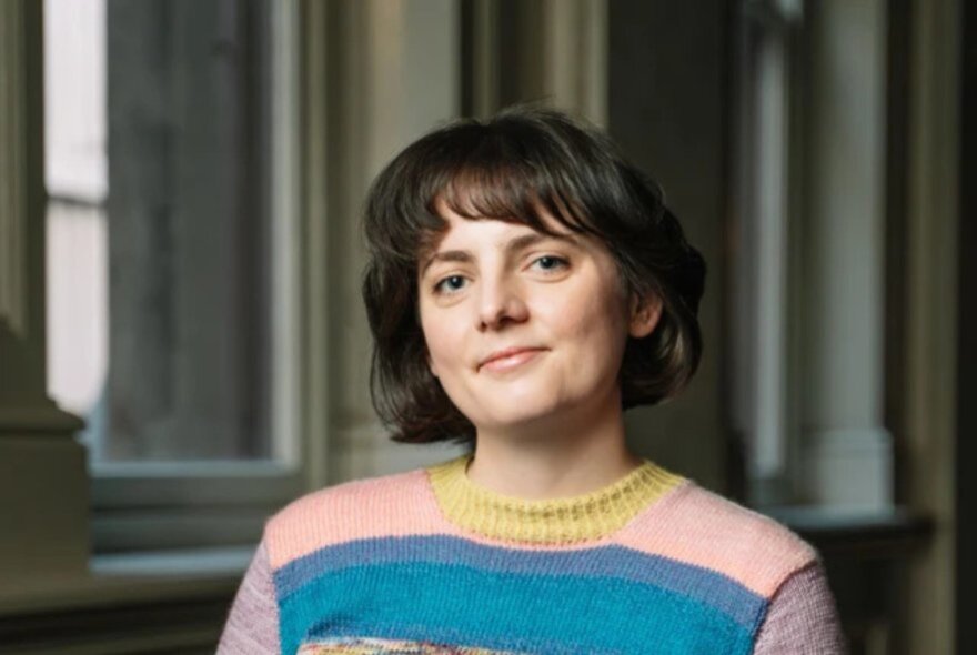 Portrait of a subtly smiling young woman with short brown hair, wearing a pink and blue striped knitted jumper.