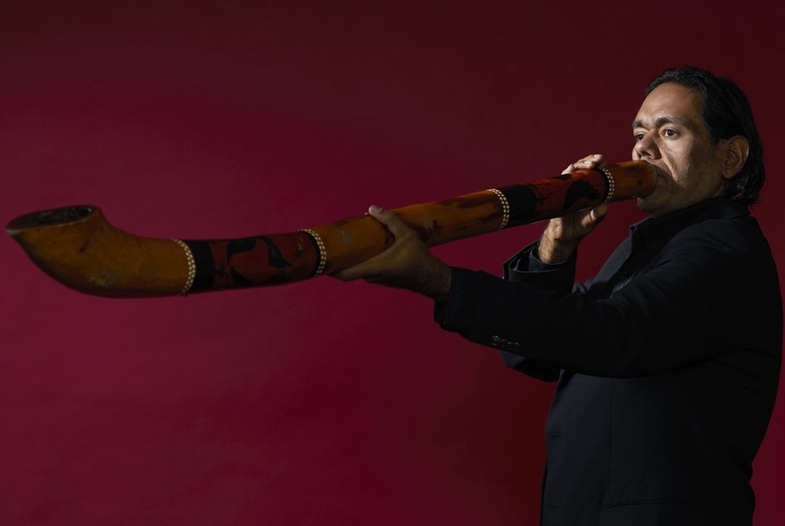 A man playing a didgeridoo, holding it out straight in front of him, against a deep red background.