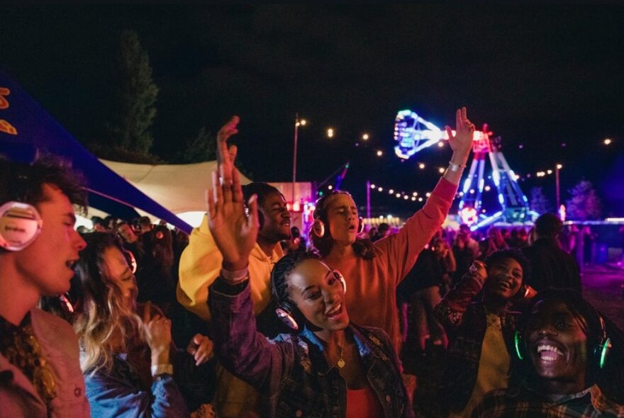 A crowd of people dancing and enjoying themselves in an open air space at night, all wearing headphones over their ears.