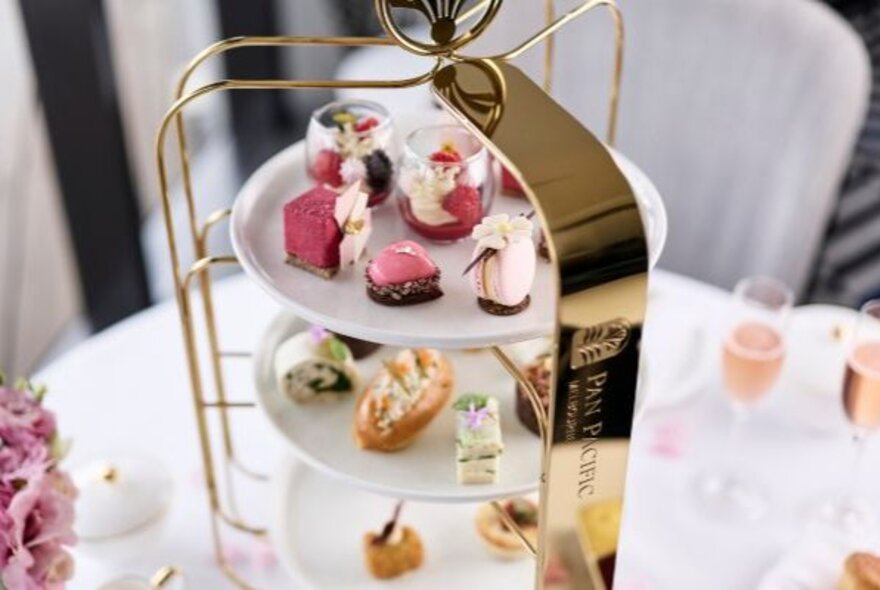A three-tiered cake stand on a white table with pink champagne. 