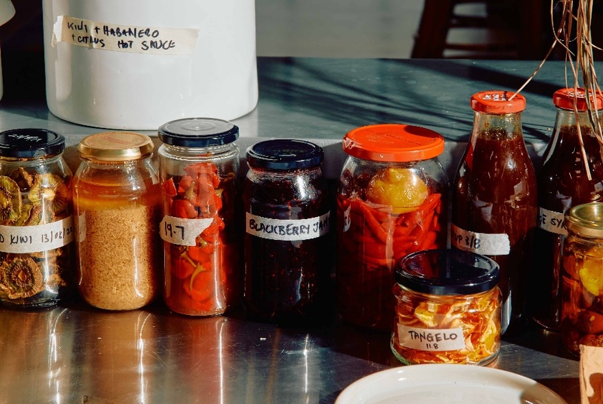 Clear glass jars of DIY fermented and pickled produce on a cafe's stainless steel kitchen bench.