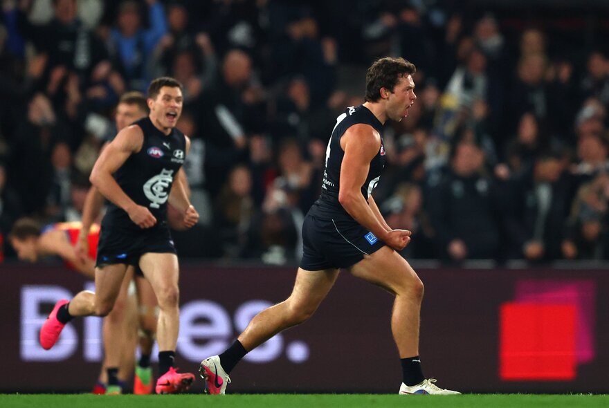 Carlton players celebrate while running on the field with a blurred crowd behind. 
