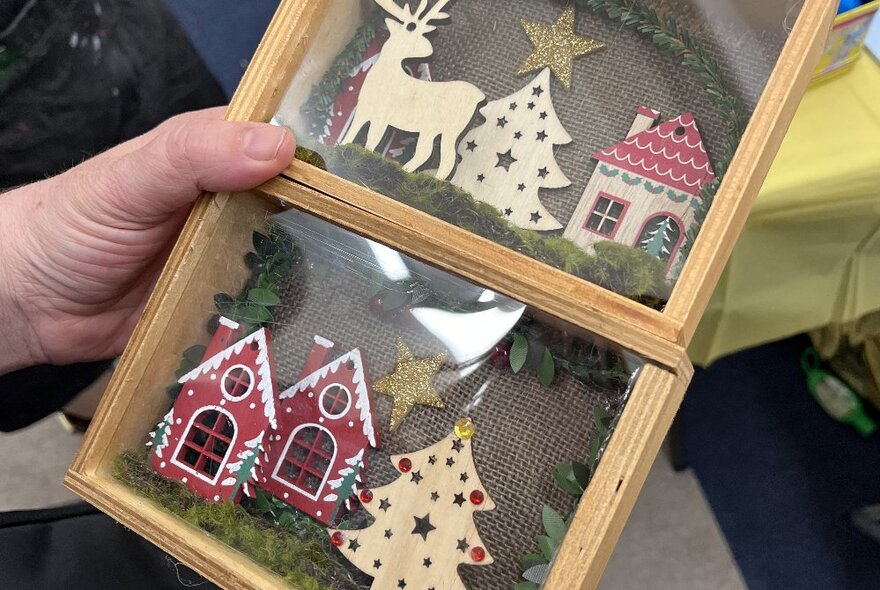 A hand holding a diorama with two frames featuring Christmas trees, reindeer and gingerbread houses. 