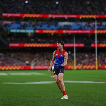 A Melbourne AFL football player on the ground during a match, the goal posts and a blurred crowd of fans in the background.
