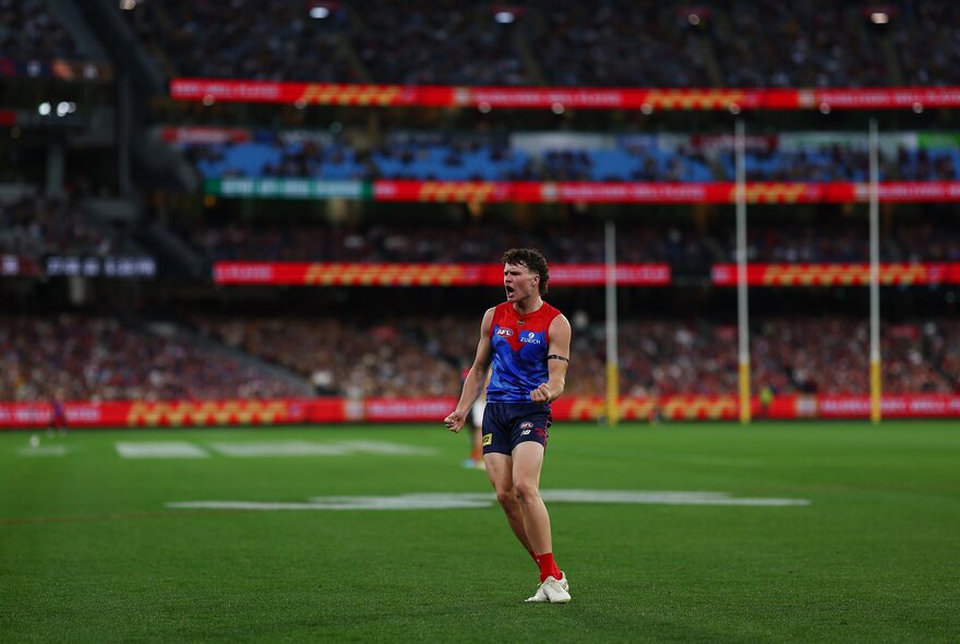 A Melbourne AFL football player on the ground during a match, the goal posts and a blurred crowd of fans in the background.