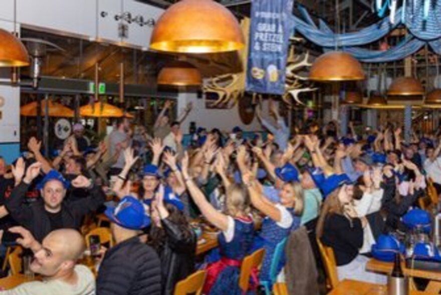 A packed crowd of people cheering with their hands raised inside a lively Bavarian-style beer hall during Oktoberfest celebratons,