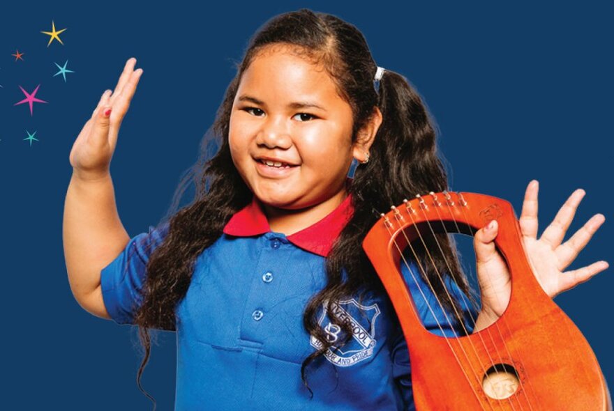 Primary school aged child, smiling and holding a stringed musical instrument, with both her hands raised in the air.