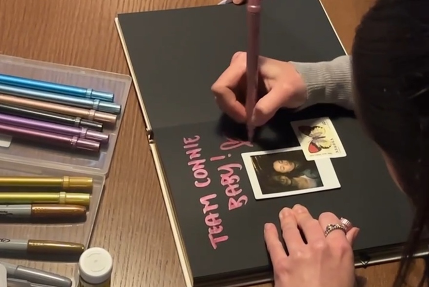 A woman using paint pens to write in a notebook with black pages and a polaroid picture pasted onto the page.