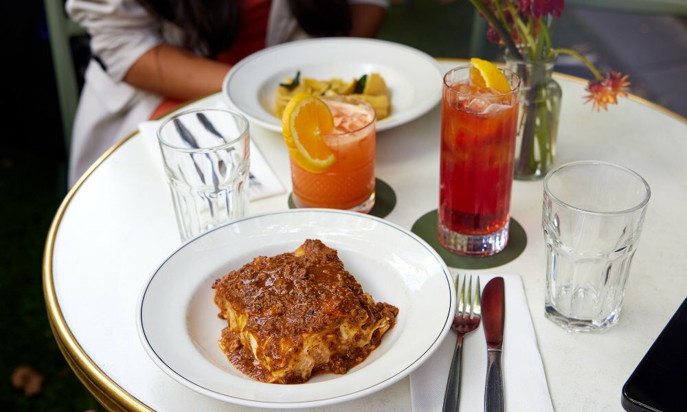 A slice of saucy lasagne with cocktails on a restaurant table.