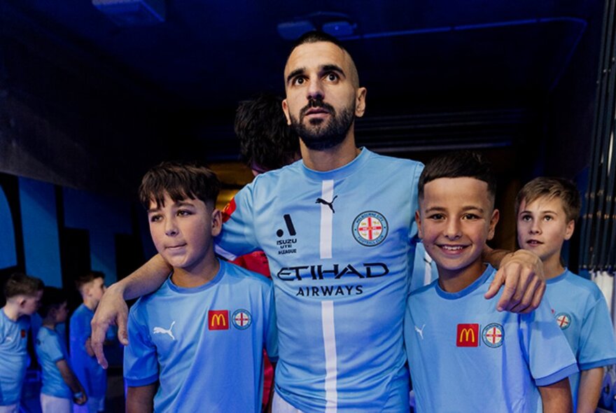 Soccer player in light-blue strip with his arms around two young mascots.