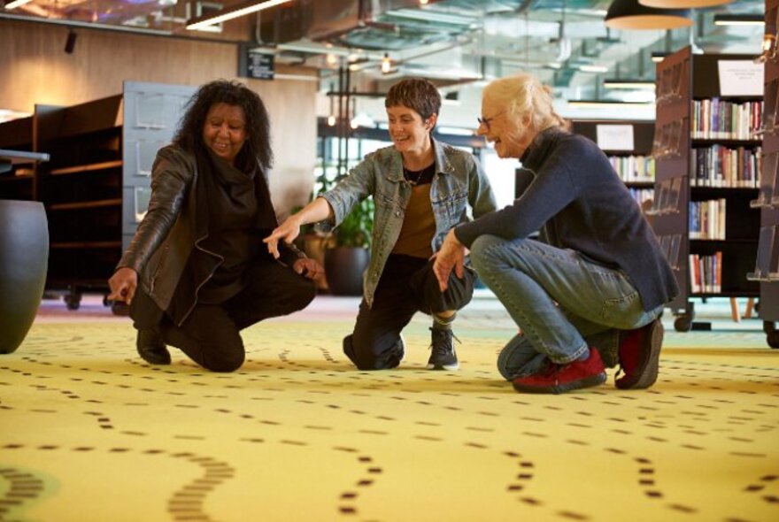 Three people crouching down on a patterned carpet in a library setting, pointing at the ground.