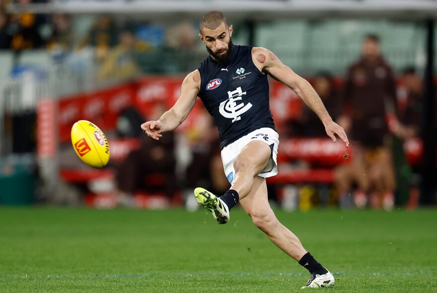 A Carlton AFL player running on the field about to kick a yellow football, with a blurred stadium crowd behind him.