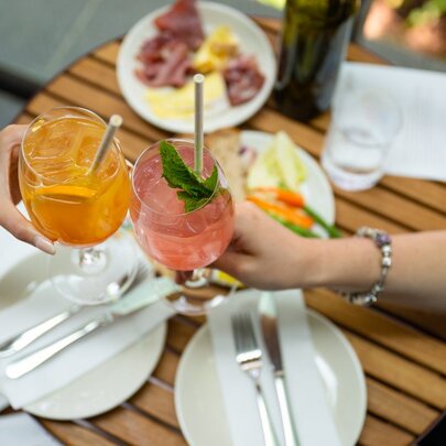 Two people clink glasses of orange and pink cocktails over a wooden table set with plates of charcuterie, vegetables, and cutlery.