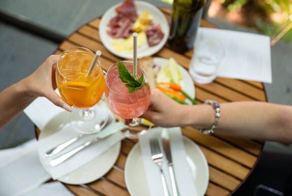 Two people clink glasses of orange and pink cocktails over a wooden table set with plates of charcuterie, vegetables, and cutlery.