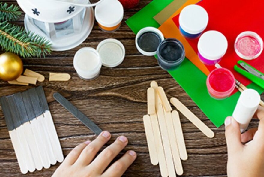 Overhead view of child's hands at a craft table with small pots of paint, a glue stick, and wooden ice-cream sticks.