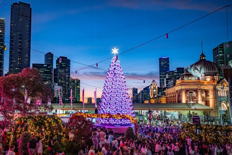 A giant illuminated purple Christmas Tree at Federation Square, surrounded by people and view of Flinders St Station and city buildings in the background.
