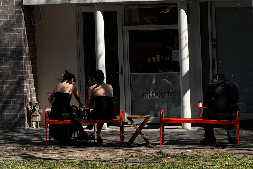 The back of people sitting on red benches in front of a small cafe.