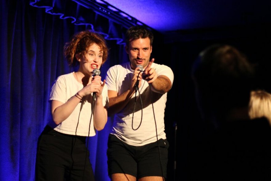 Two comedians performing on a small stage in front of an audience, each holding microphones.