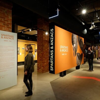 Installation view of the Spartans & Anzacs exhibition at the Shrine of Remembrance, with people walking around the space, reading panels and looking at displays.