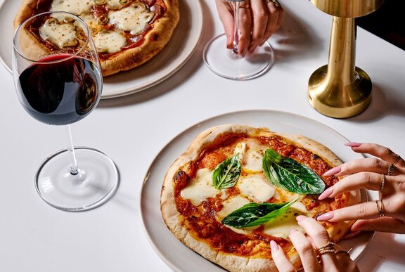 A woman's hands with manicured nails holding the edges of a plated woodfired pizza, a glass of red wine on the table next to it.