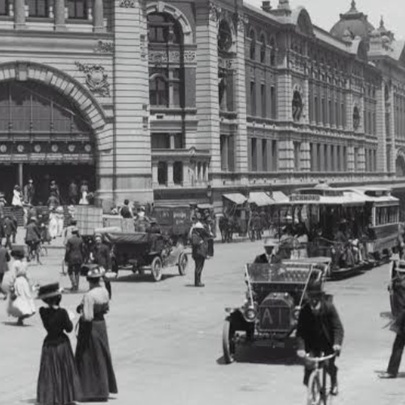 Black and white historical photograph of a busy street intersection in front of the grand, multi-story Flinders Street railway station building in Melbourne.