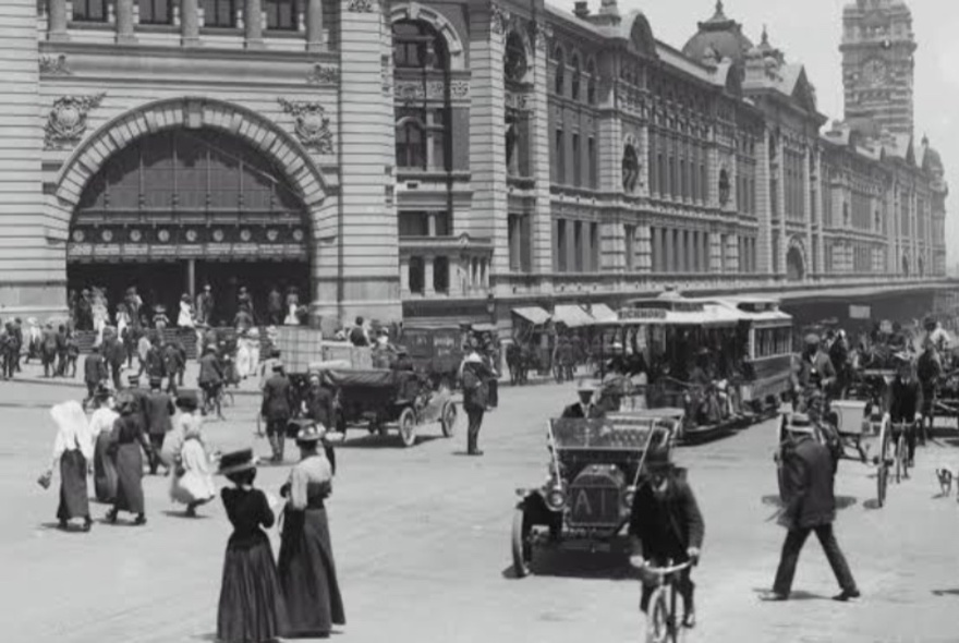 Black and white historical photograph of a busy street intersection in front of the grand, multi-story Flinders Street railway station building in Melbourne.
